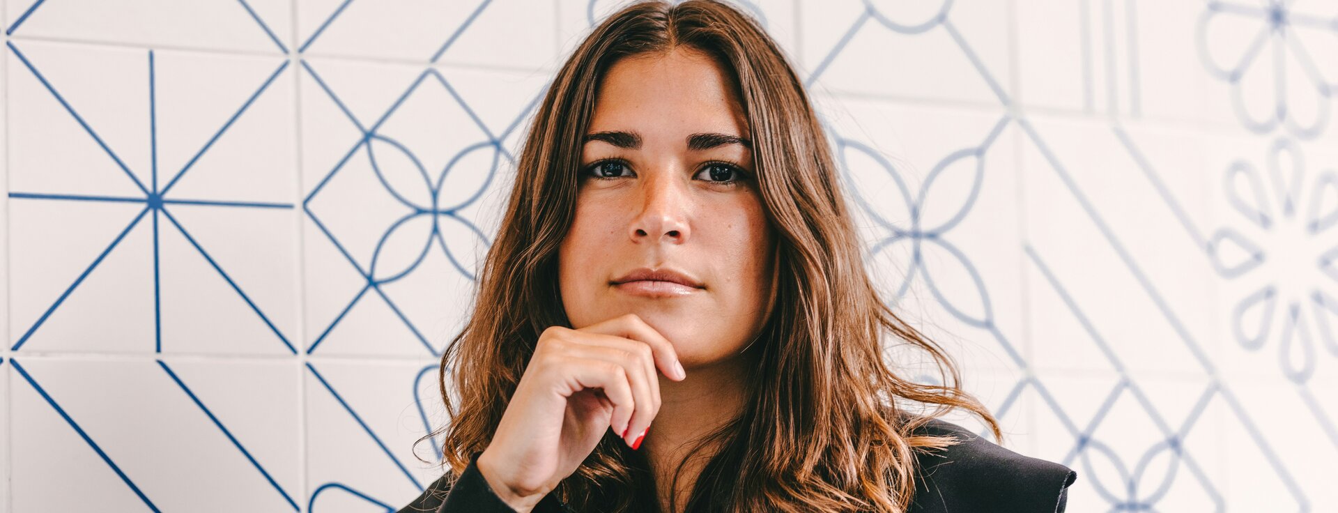 Junge Frau mit schwarzem Blazer, die vor einer Wand mit weiß-blauen Fliesen steht und nachdenkt  | © Ruben Ramirez, Unsplash